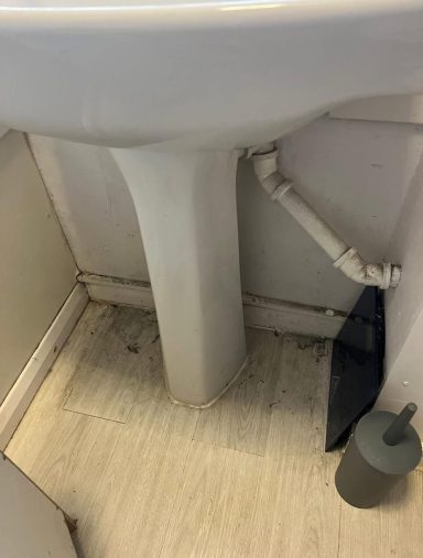 Close-up of a bathroom sink pedestal with visible plumbing and floor in a compact space.