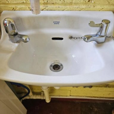 White sink with two chrome taps and a central drain, mounted on a yellow wall.