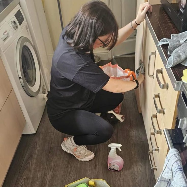 Hayley 2 Person cleaning in a kitchen, crouching near a cupboard with cleaning supplies nearby.