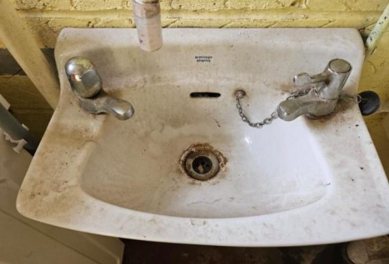 A dirty, old washbasin with worn taps and a stained surface.
